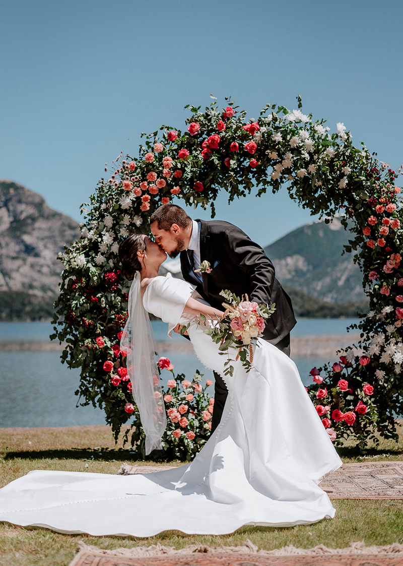 El novio besa con pasión a la novia, que luce un hermoso vestido de larga cola frente al altar de flores, en la celebración de su boda en la magnífica Bariloche, Patagonia, Argentina. Dupla Planners lo hizo posible.